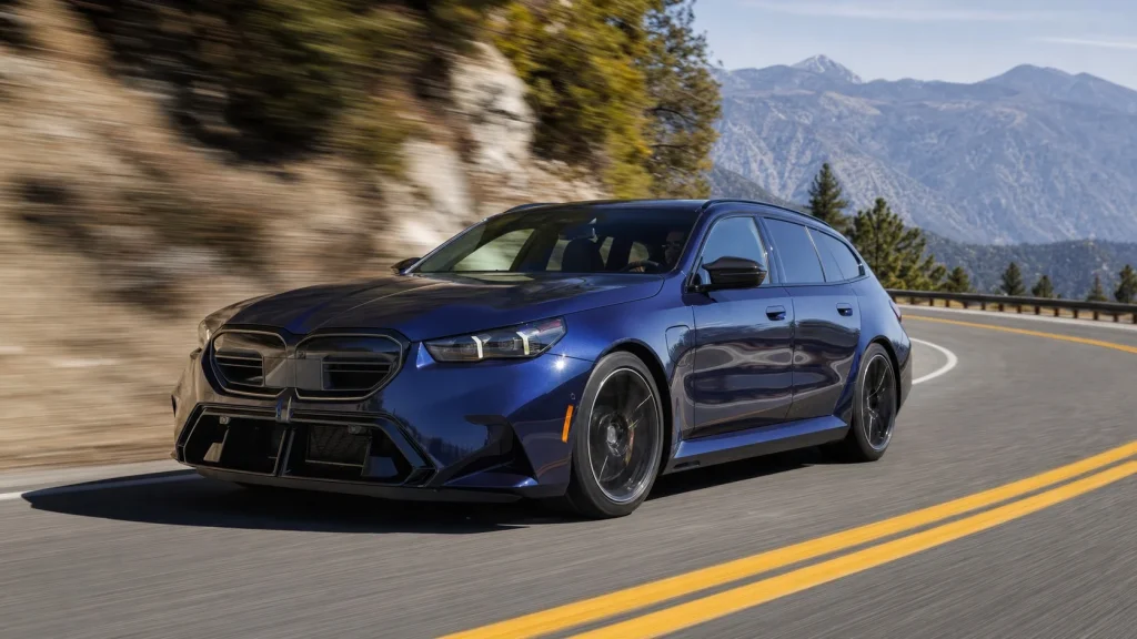 A 2026 BMW M5 Touring cornering at high speed on a scenic mountain road.